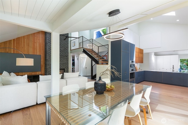 dining room featuring wooden walls, light wood finished floors, a fireplace, high vaulted ceiling, and stairs
