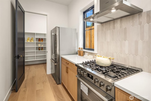 kitchen with backsplash, light countertops, light wood finished floors, stainless steel appliances, and wall chimney exhaust hood