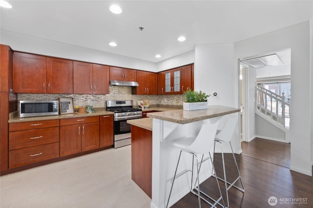 kitchen with a kitchen breakfast bar, tasteful backsplash, under cabinet range hood, glass insert cabinets, and stainless steel appliances
