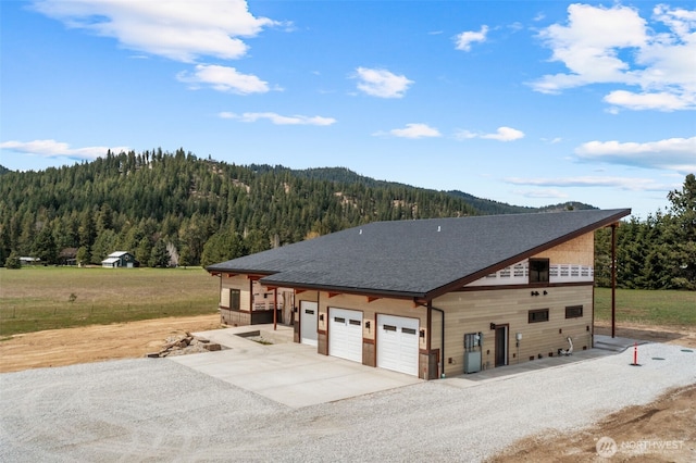 view of property exterior featuring roof with shingles, a garage, a mountain view, gravel driveway, and a wooded view
