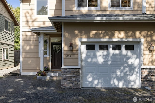 doorway to property featuring an attached garage, a shingled roof, stone siding, and gravel driveway