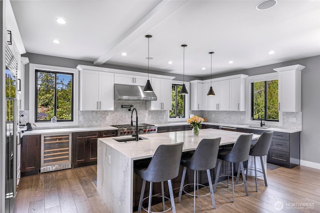 kitchen with wine cooler, a kitchen island with sink, under cabinet range hood, a sink, and backsplash