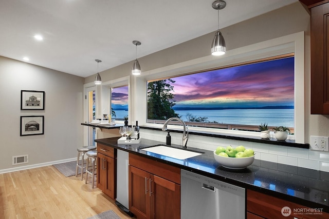 kitchen with dishwasher, a sink, brown cabinets, dark stone counters, and light wood-style flooring