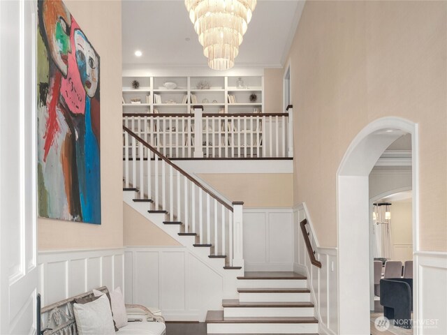 staircase with wainscoting, ornamental molding, a decorative wall, and a notable chandelier