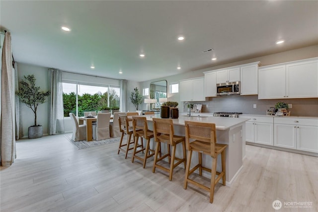 kitchen with decorative backsplash, stainless steel microwave, an island with sink, and light wood finished floors