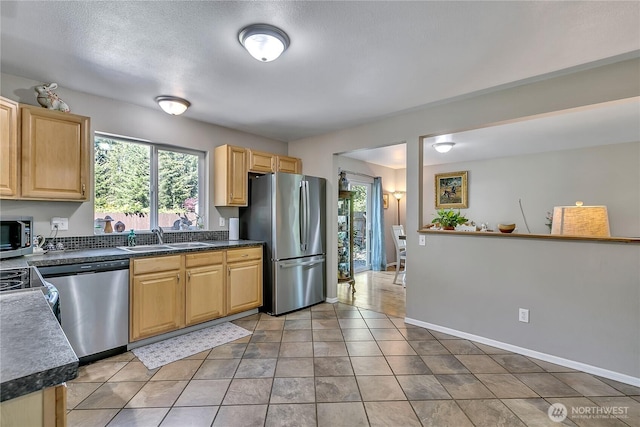 kitchen with appliances with stainless steel finishes, light brown cabinetry, dark countertops, a sink, and baseboards