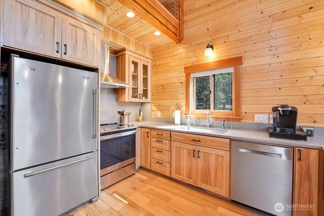 kitchen with wall chimney range hood, wood walls, a sink, light wood finished floors, and stainless steel appliances