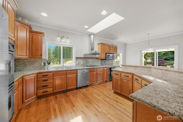 kitchen with wall chimney range hood, a skylight, appliances with stainless steel finishes, a sink, and light wood-style floors