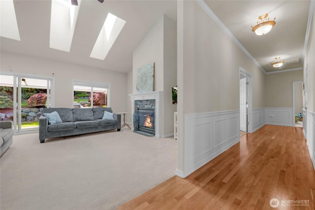 living area featuring a stone fireplace, a skylight, light wood-type flooring, a wainscoted wall, and ornamental molding