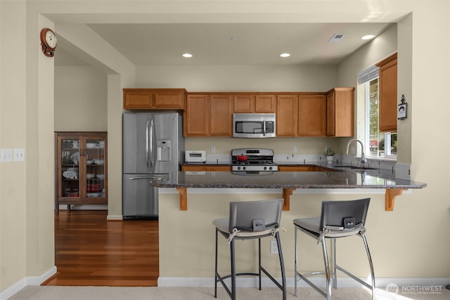 kitchen featuring appliances with stainless steel finishes, a sink, dark stone countertops, brown cabinetry, and recessed lighting