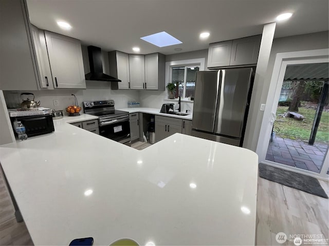 kitchen with wall chimney range hood, appliances with stainless steel finishes, a sink, a skylight, and gray cabinetry