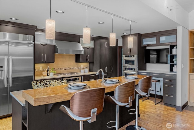 kitchen with a breakfast bar area, wall chimney range hood, light countertops, light wood-style flooring, and stainless steel appliances