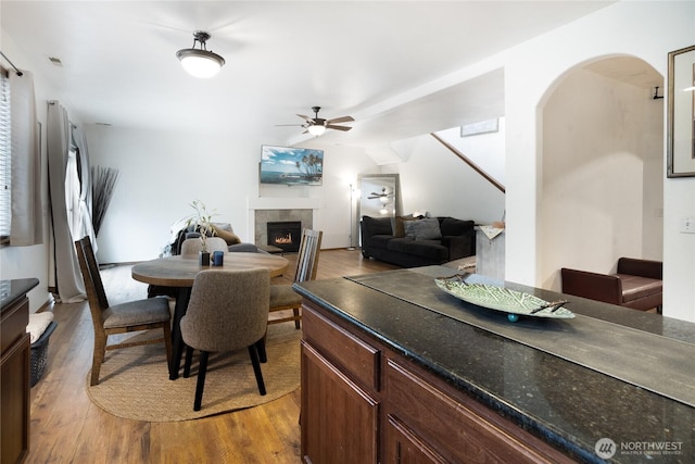 dining area featuring a ceiling fan, wood finished floors, arched walkways, and a tiled fireplace