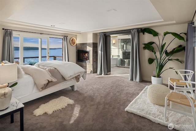 bedroom featuring access to exterior, a water view, carpet flooring, a tile fireplace, and a tray ceiling