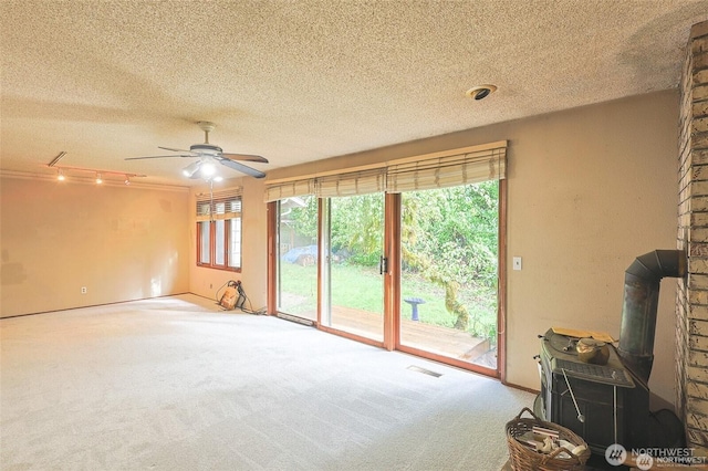 unfurnished living room with ceiling fan, a wood stove, carpet floors, a textured ceiling, and visible vents
