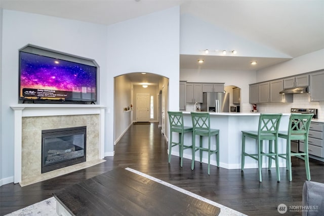 kitchen with gray cabinetry, stainless steel fridge, under cabinet range hood, a kitchen bar, and dark wood finished floors