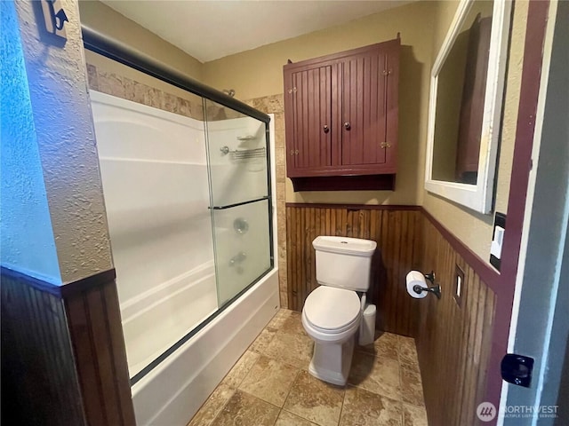 full bathroom with toilet, enclosed tub / shower combo, wainscoting, and stone finish floor