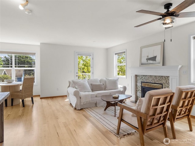 living room with light wood finished floors, a tiled fireplace, a ceiling fan, and baseboards