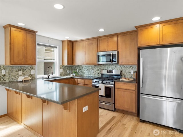 kitchen featuring stainless steel appliances, a sink, a peninsula, light wood finished floors, and brown cabinets