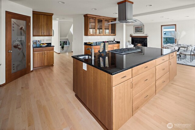 kitchen with a sink, stainless steel gas cooktop, light wood-style floors, a lit fireplace, and a kitchen island with sink
