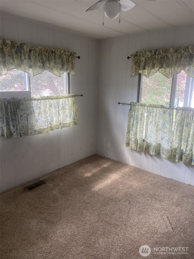carpeted spare room featuring a healthy amount of sunlight, a ceiling fan, wooden walls, and visible vents
