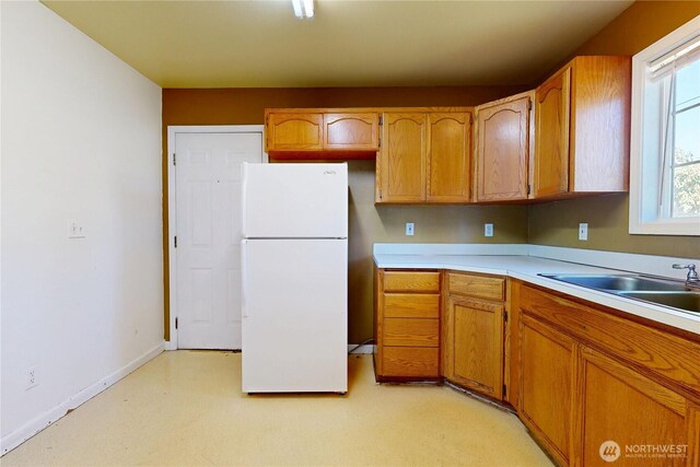 kitchen featuring freestanding refrigerator, light countertops, a sink, baseboards, and brown cabinetry