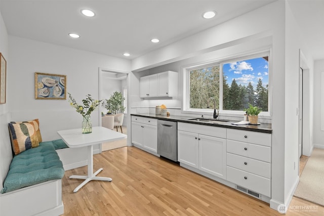 kitchen featuring stainless steel dishwasher, dark countertops, white cabinetry, a sink, and light wood-style flooring