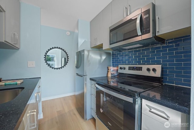 kitchen featuring stainless steel appliances, light wood finished floors, tasteful backsplash, dark stone counters, and a sink