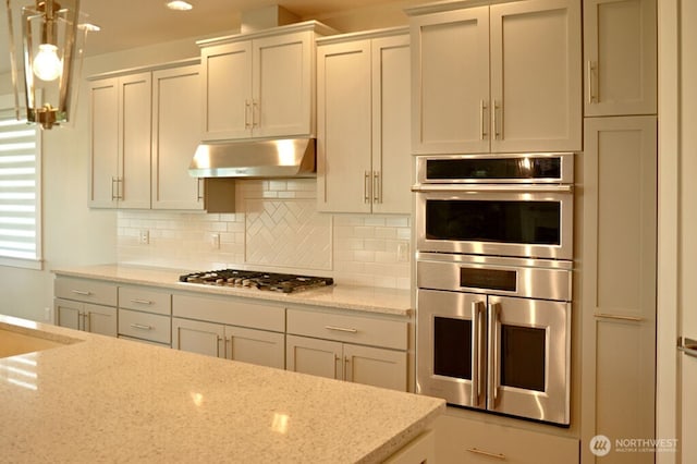 kitchen with under cabinet range hood, appliances with stainless steel finishes, light stone counters, backsplash, and hanging light fixtures