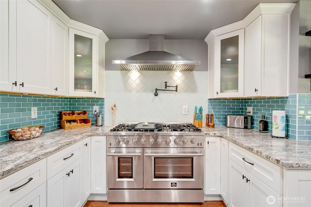 kitchen featuring range with two ovens, wall chimney exhaust hood, tasteful backsplash, and white cabinetry