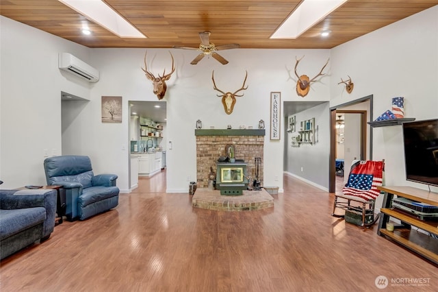 living area featuring a wall mounted AC, wood ceiling, a ceiling fan, a skylight, and a wood stove