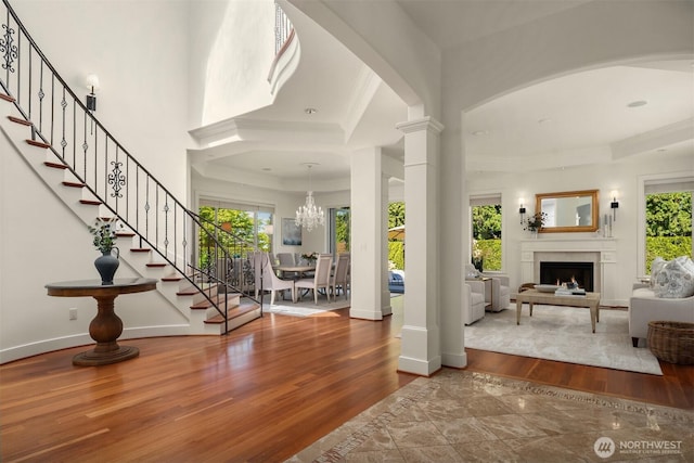 foyer with stairway, wood finished floors, a chandelier, a warm lit fireplace, and decorative columns