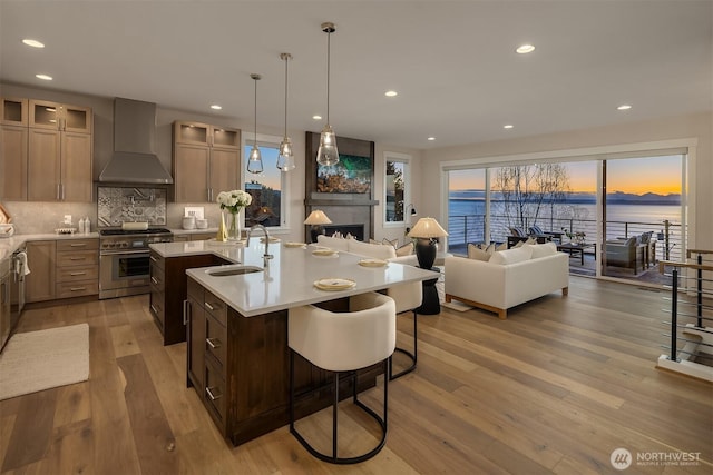 kitchen with high end stainless steel range oven, a sink, wall chimney exhaust hood, a center island with sink, and light countertops