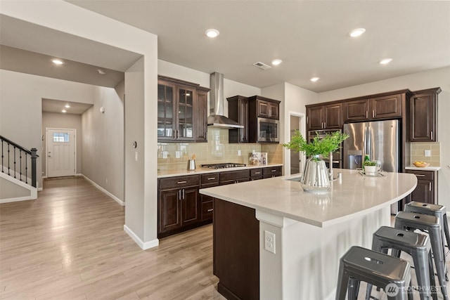 kitchen with wall chimney range hood, appliances with stainless steel finishes, a kitchen bar, light countertops, and decorative backsplash