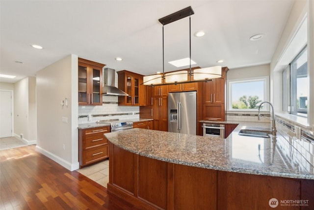 kitchen with wall chimney exhaust hood, stainless steel appliances, a sink, brown cabinets, and light stone countertops