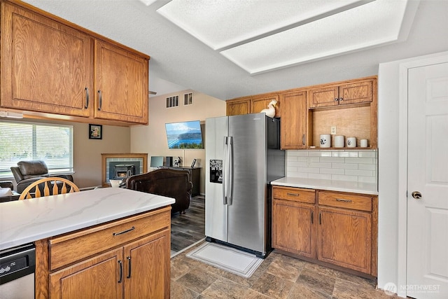 kitchen with stainless steel fridge, brown cabinetry, decorative backsplash, stone finish floors, and lofted ceiling
