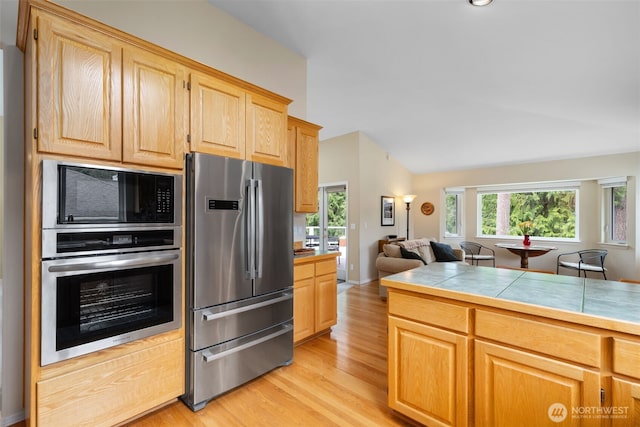 kitchen featuring appliances with stainless steel finishes, light wood finished floors, tile counters, light brown cabinetry, and vaulted ceiling