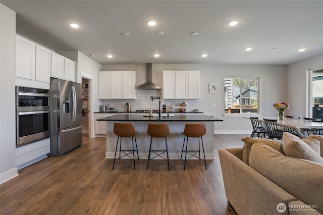 kitchen featuring appliances with stainless steel finishes, wall chimney range hood, dark countertops, a kitchen bar, and decorative backsplash
