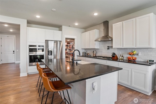 kitchen featuring wall chimney exhaust hood, stainless steel appliances, a sink, tasteful backsplash, and light wood finished floors