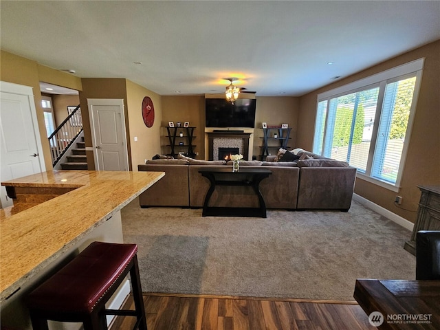 carpeted living room with a ceiling fan, a warm lit fireplace, baseboards, wood finished floors, and stairway