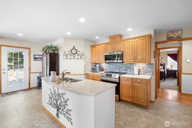 kitchen featuring stainless steel appliances, a sink, decorative backsplash, an island with sink, and light stone counters