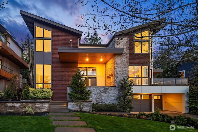 back of property at dusk with a lawn, a garage, stone siding, and a balcony