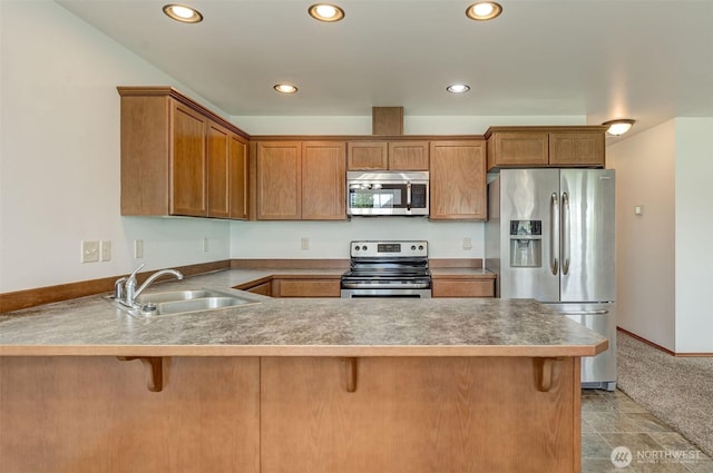 kitchen featuring stainless steel appliances, a sink, a peninsula, recessed lighting, and brown cabinetry