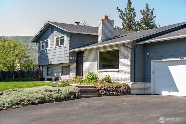split level home featuring aphalt driveway, fence, an attached garage, brick siding, and a chimney