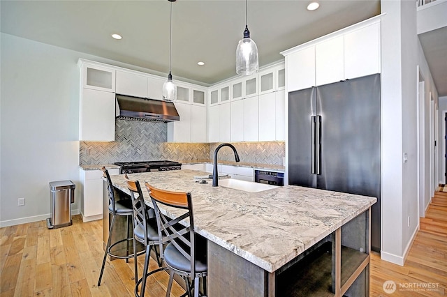 kitchen with a sink, light wood-style flooring, high end refrigerator, decorative backsplash, and under cabinet range hood