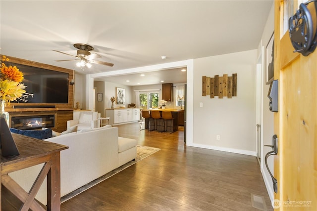 living area featuring dark wood-style flooring, a ceiling fan, a glass covered fireplace, baseboards, and recessed lighting