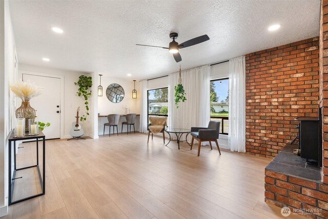 living room featuring a ceiling fan, brick wall, a textured ceiling, light wood-style flooring, and a wood stove