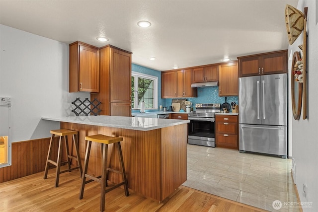 kitchen with appliances with stainless steel finishes, brown cabinets, a peninsula, light countertops, and a wainscoted wall