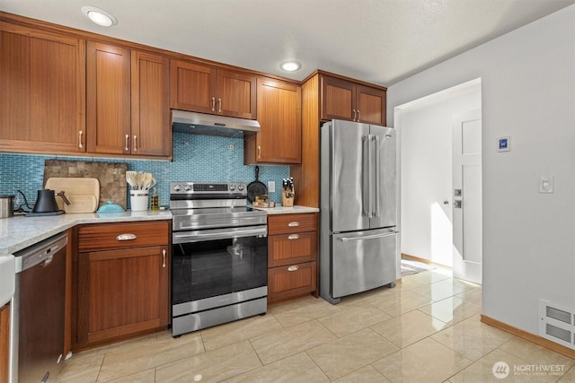 kitchen with stainless steel appliances, under cabinet range hood, brown cabinets, baseboards, and tasteful backsplash