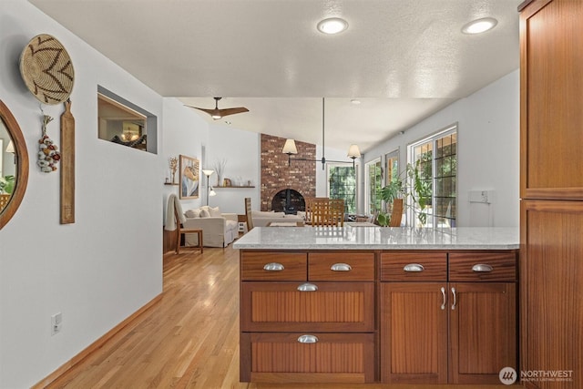 kitchen featuring a brick fireplace, light wood-style floors, brown cabinetry, light stone counters, and a ceiling fan